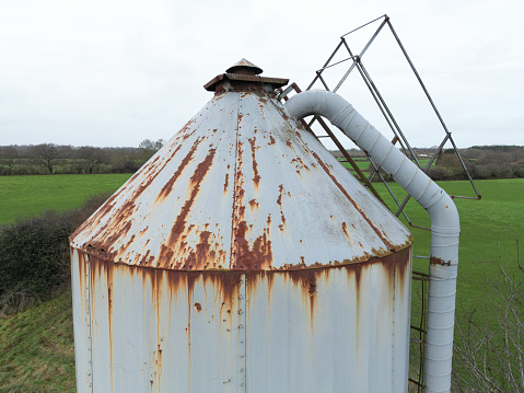 Large, metal grain feed silo showing a  poor state of repair. Surrounding farm buildings are seen belonging to a British dairy farm.