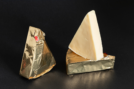 Triangular cream cheeses wrapped in golden aluminium foil and without packaging on a black background. Portioned triangular cheeses.