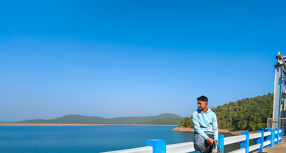 A young man sitting on the bridge and looking at blue river landscape view, lake with blue sky scenery. Holiday concept