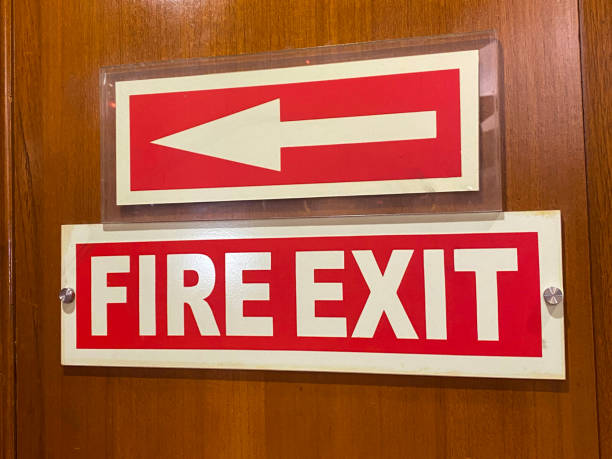 Full frame image of rectangular red and white 'Fire Exit' sign on wooden wall panelling with white arrow on red rectangular direction advisory sign, focus on foreground Stock photo showing close-up view of a wooden wall panel with a rectangular red and white 'Fire Exit' sign screwed to it.  A rectangular, red and white arrow sign is fixed above the 'Fire Exit' sign. The sign advices of the direction to the emergency fire door. door fire exit sign swinging doors fire door stock pictures, royalty-free photos & images