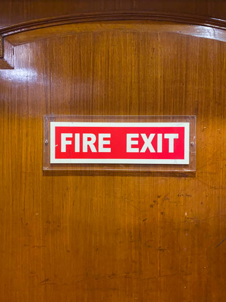 Close-up image of rectangular red and white 'Fire Exit' sign on emergency fireproof door, focus on foreground Stock photo showing close-up view of a wooden emergency fire door with a rectangular red and white 'Fire Exit' sign screwed to it. door fire exit sign swinging doors fire door stock pictures, royalty-free photos & images