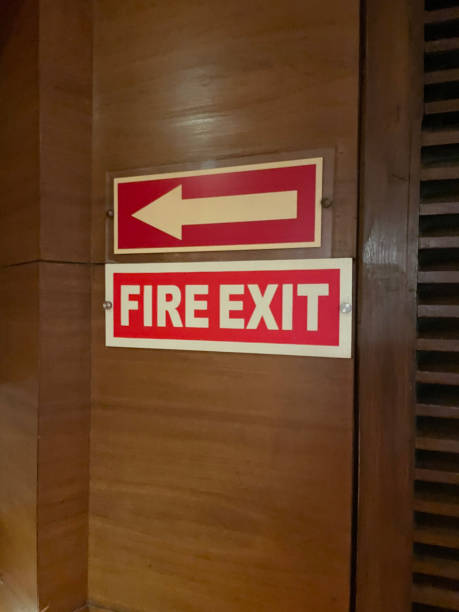 Image of rectangular red and white 'Fire Exit' sign on wooden wall panelling with white arrow on red rectangular direction advisory sign, focus on foreground Stock photo showing close-up view of a wooden wall panel with a rectangular red and white 'Fire Exit' sign screwed to it.  A rectangular, red and white arrow sign is fixed above the 'Fire Exit' sign. The sign advices of the direction to the emergency fire door. door fire exit sign swinging doors fire door stock pictures, royalty-free photos & images