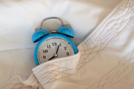 Stock photo showing close-up view of retro, double bell, analogue alarm clock tucked up for bedtime on pillow and under bedding.