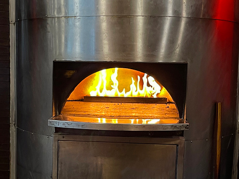 Stock photo showing close-up view of metal, pizza oven at Italian pizzeria restaurant, with burning flame and glowing embers.