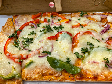 Stock photo showing close-up, elevated view of cardboard takeaway pizza box containing a sliced vegetarian pizza, topped with a rich tomato sauce, melted buffalo mozzarella and garnished with red, yellow and green bell pepper and red onion.