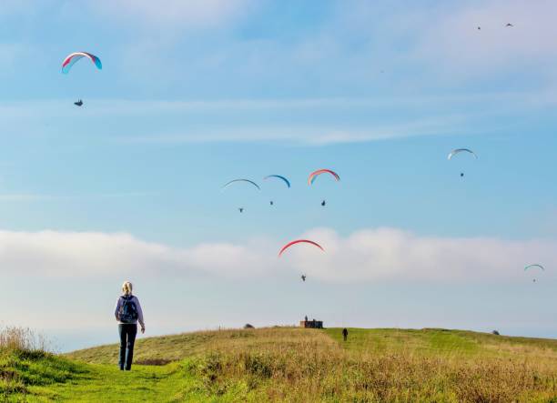 A hiker approaching Paragliders flying over Beachy Head, East Sussex, UK A female hiker walks towards a colourful cloud of Paragliders flying above Beachy Head, near Eastbourne, East Sussex, UK.  This is a well known beauty spot in the South Downs National Park. eastbourne pier photos stock pictures, royalty-free photos & images