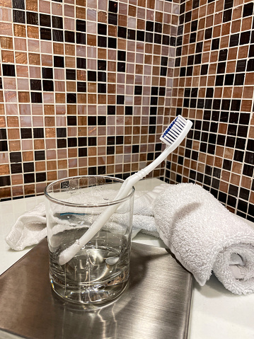 Stock photo showing a hotel bathroom featuring a white toothbrush in a drinking glass besides a rolled up towel on a marble bathroom counter.