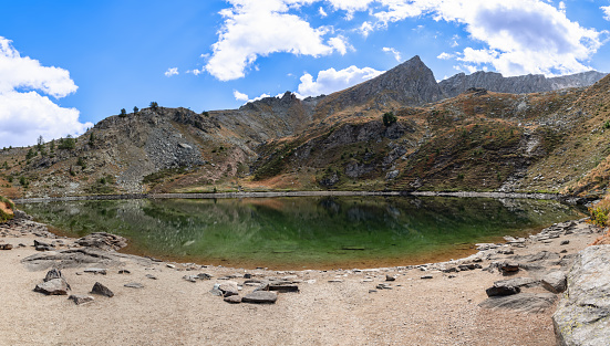 Crystal-clear water of Lago di Loie at 2354m with panoramic view of surrounding autumn cliffs and peaks under blue sky with white clouds, Aosta Valley, Italy