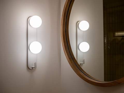 Modern mounted wall light lamp near the round shape wooden mirror on white wall background in the bathroom. Lighting, ball shape decoration on the corner of the bathroom wall in the hotel.