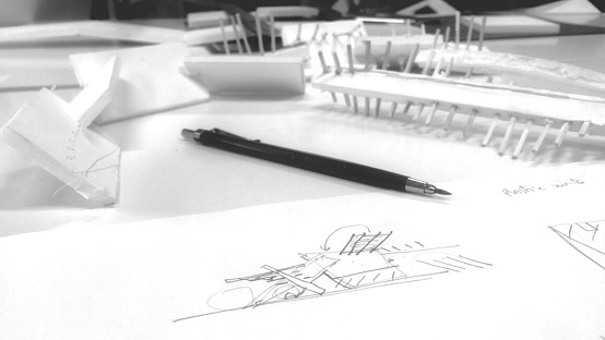 Pencil on architectural sketches and physical model on desk. Black and white image of architect's working desk with building drawing and 3d model.