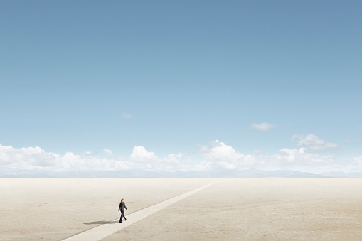 A lone woman takes a long walk on a path toward the horizon on a sunny day across a barren desert floor.