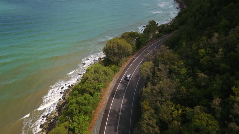 Car driving along road at tropical Cape Tribulation at Daintree Rainforest, Australia. Beautiful aerial drone from above. Clear blue water white sandy beach and jungle palm trees at the seaside. Perfect island adventure. Moving modern automobile 4K UHD.