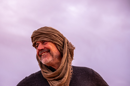 Portrait of a smiling senior man with tagelmust on his head and the sky in the background. The tagelmust, cheich, cheche or litham is a cotton turban-like garment. It is mainly worn by Tuareg Berber men.