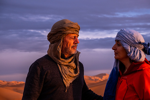 Portrait of a senior couple with tagelmust on his head and the sky and desert dunes in the background. The tagelmust, cheich, cheche or litham is a cotton turban-like garment. It is mainly worn by Tuareg Berber men.