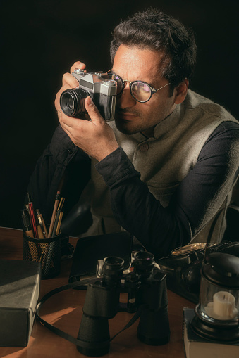 An Asian/Indian mid-adult man in the role of a photojournalist of the 70s holds an SLR film camera sitting on the office table reflecting the setting of the 70s.