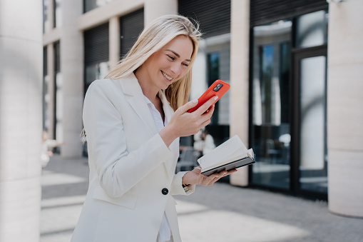 Amazed businesswoman in whit suit holding phone toothy smiling, happy received great news, walking by the street. Attractive Italian entrepreneur making video call during a break. Surprised female