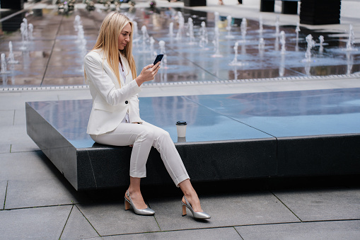 Gorgeous Blonde caucasian young woman sitting outside, having coffee break, dressed in white suit looking up, holding phone preparing to make an important call. Purposeful businesswoman thinks