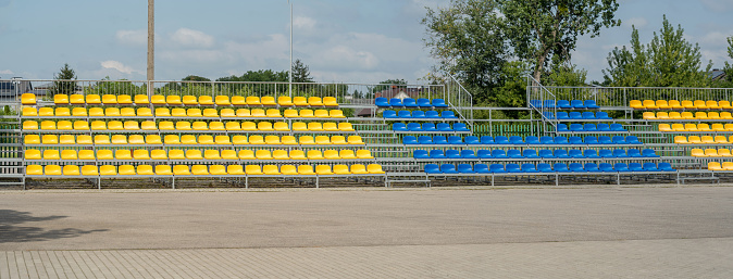Simple outdoor vacant empty stadium stands wide panorama shot, nobody, no people, small downtown sports arena outdoors venue without people. Lots of yellow and blue stadium stand chairs, daytime