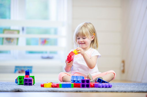 A sweet young blond haired girl sits on the floor in a home daycare as she plays with colorful blocks alone.  She is dressed casually and is smiling as she enjoys the free time.