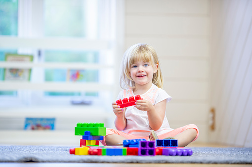 A sweet young blond haired girl sits on the floor in a home daycare as she plays with colorful blocks alone.  She is dressed casually and is smiling as she enjoys the free time.