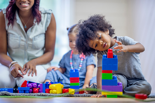 A female home daycare provider of African decent, sits on the floor with some toddlers as they play with blocks together.  They are each dressed casually and are smiling as they enjoy the free play time.