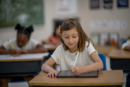 A female elementary student sits at her desk with a tablet in front of her as she works away diligently on the device.  She is dressed casually and her peers can be seen working away in the background.
