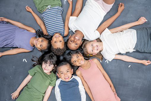 A large group of elementary students lay on the floor in a circle with their heads together, as they pose a for a portrait.  They are each dressed casually and are smiling in this aerial view photo.