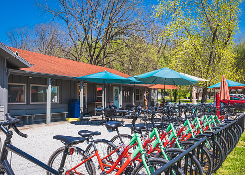 View of patio of Midwestern restaurant and bike shop on nice sunny day; row of bicycles in foreground; café building, umbrellas and several people behind them