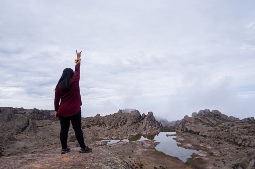 latin woman with her back turned and her arm raised with the sign of the metal, standing on the edge of a cliff with a lagoon in the background.