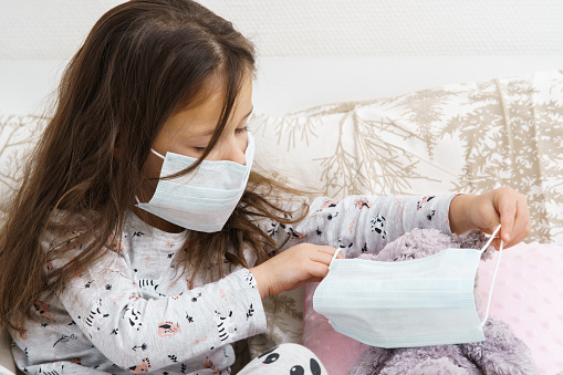 Sick little girl in sleepwear sitting on couch and playing with teddy bear. Portrait of child, preschooler. Kid and toy wear medical masks on face. Patient treatment, protective measures, disease.