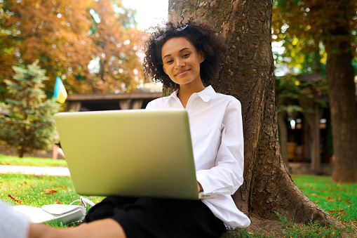Work remotely. Dark-haired girl sitting under the tree with a laptop