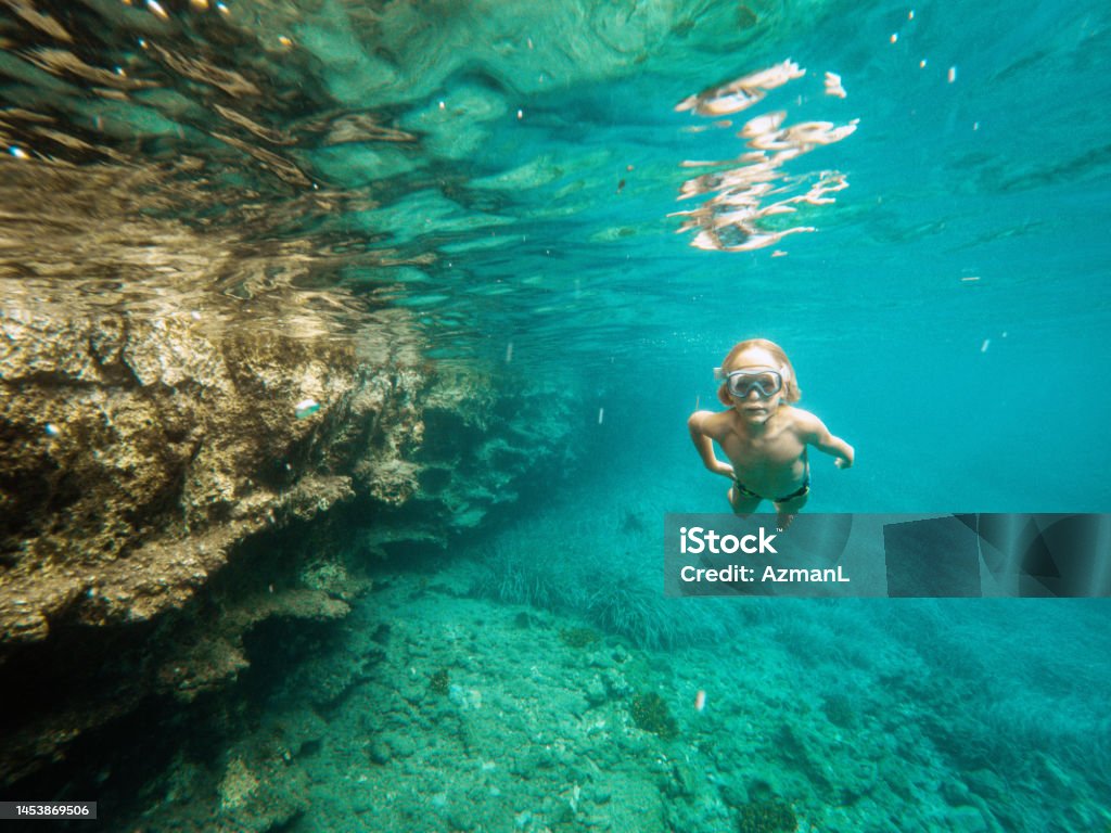 Small Caucasian Boy Diving Underwater And Exploring In The Sea Happy boy having a wonderful underwater time in a national park. He is swimming, diving and exploring in nature. He is wearing goggles and looking at the camera. 6-7 Years Stock Photo