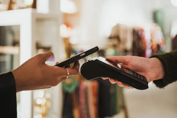 Close up shot of young woman paying contactless on a POS in a shop. She is using her phone.