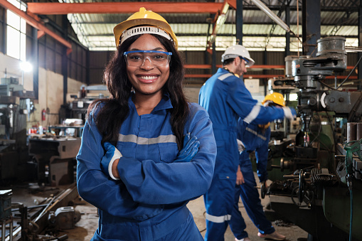 Portrait of professional Black female worker in protective safety uniform and helmet looking at camera, arms crossed and smiling, with engineers team behind her in a metalwork manufacturing factory.