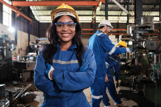 Portrait of a female industrial worker in a metalwork manufacturing factory. Portrait of professional Black female worker in protective safety uniform and helmet looking at camera, arms crossed and smiling, with engineers team behind her in a metalwork manufacturing factory. manufacturing occupation stock pictures, royalty-free photos & images