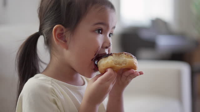 Asian little girl dirty mouth eating a donut at home in the living room while watching TV
