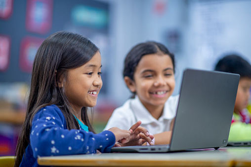 Two elementary students sit at a desk as they work together on a laptop during a computer lab.  They are each dressed casually and smiling as they focus on the screen.