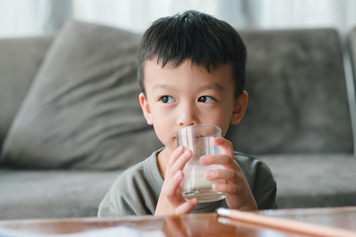 Close up of happy Asian little cute kid or child boy hand holding milk glass and drinking milk during sitting on the sofa at home before study. Daily life health care Medicine food