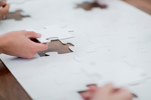 A small group of adults are seen putting together a blank, all white, jigsaw puzzle while seated around a wooden table.  Only the adults hands are seen as they each put in one last piece to finish the puzzle.