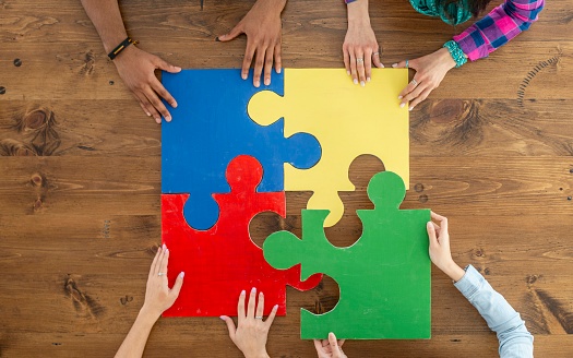 A small group of four adults are seen each putting in a different colored puzzle piece to finish the small 4 piece jigsaw puzzle.  Only the adults hands are seen as they each fit their piece in.