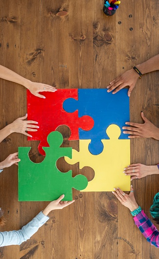 A small group of four adults are seen each putting in a different colored puzzle piece to finish the small 4 piece jigsaw puzzle.  Only the adults hands are seen as they each fit their piece in.