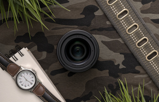 Overhead flat lay of a 30mm f1.4 digital camera lens surrounded by personal items on a camouflage background. Ideal for military, landscape, wildlife, or safari photography.