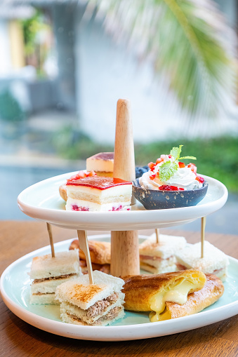 Afternoon tea with fresh cakes, pastries and sandwiches with hot tea and teapot, tropical greenery on background. Sweet breakfast in hotel. High tea desserts with sweets assortment.