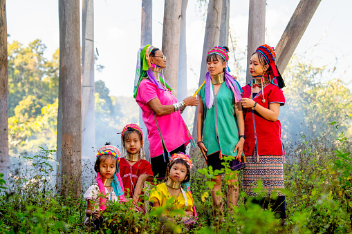 Various generation include young teen and old woman help to decorate and set hair and cloth of Long Neck Karen girl who stand in area of ceremonial courtyard of their village.