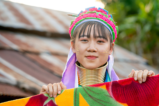 Lovely AsianLong Neck Karen girl hold multicolor blanket on clothesline and look at camera with smiling and house roof as background.