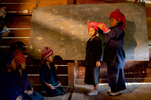 Lovely Asian hill tribe girl help to set and decorate headband of her friend in classroom with Karen alphabet show on blackboard during wait to study in school.