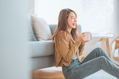 Joyful young asian female enjoying a cup of coffee while sitting on the rug beside to the sofa at home, Cosy scene, Smiling pretty woman drinking hot tea in winter.