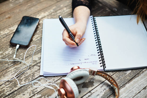 屋外のテーブルで渦巻き状のノートに書く若い女性、携帯電話とヘッドフォン - spiral notebook audio ストックフォトと画像