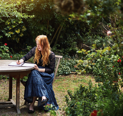 Looking like a Pre-Raphaelite painting, a woman with long flaming red hair sits outdoors at a rustic table, writing in her notebook.