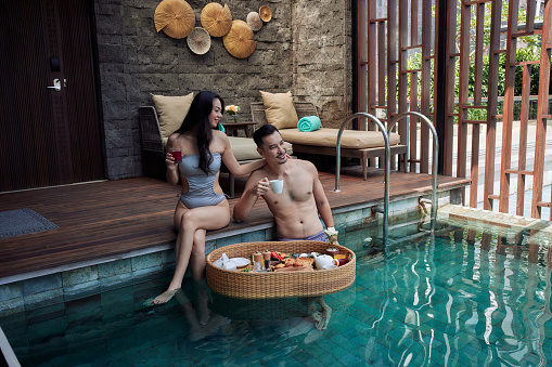 Happy Asian couple having breakfast in the pool. Floating food tray in luxury hotel. Man and woman are drinking morning coffee wearing swimsuits